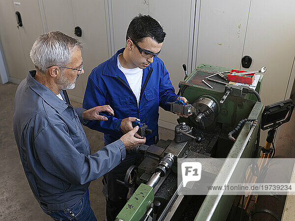 Instructor guiding apprentice using lathe machine at workshop