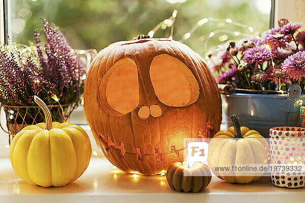 Halloween decoration of Jack O' Lantern with pumpkins and flowers near candle on window sill
