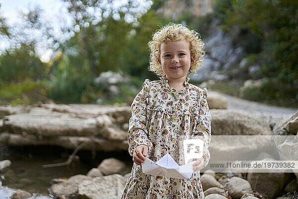 Smiling cute girl holding paper boat in front of rocks