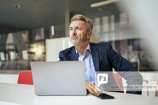 Thoughtful mature businessman sitting with smart phone and laptop at table