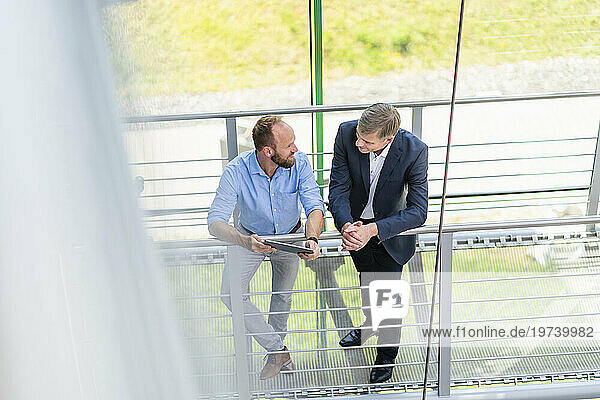 Colleagues leaning on railing in office building discussing business strategy using digital tablet