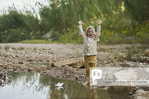 Happy girl with arms raised and paper boat in water puddle near rocks