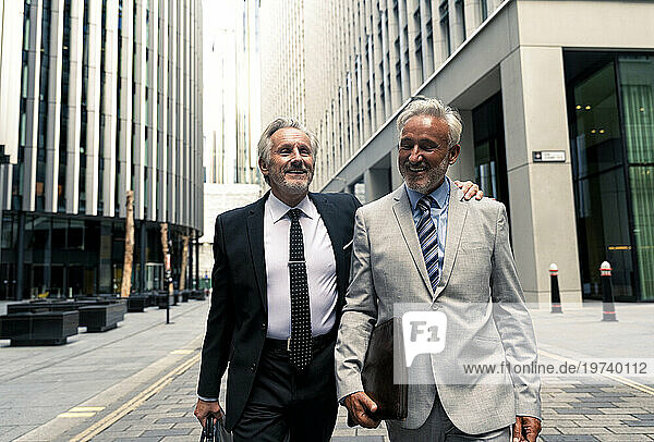 Happy senior businessmen walking and talking with each other amidst financial buildings