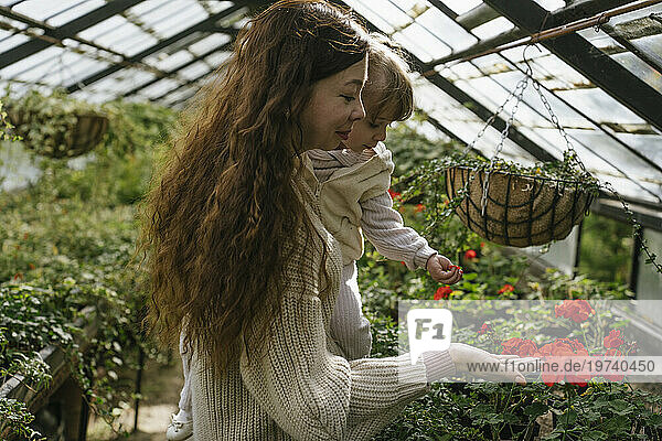 Smiling mother carrying daughter and touching flowers in greenhouse