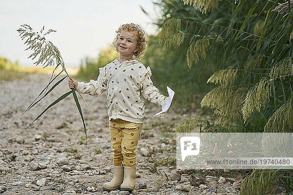 Smiling girl holding twig with paper boat and standing near plants