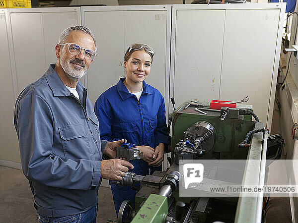 Smiling trainee standing with instructor near lathe machine at workshop