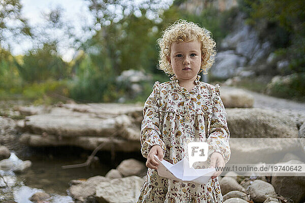 Cute girl holding paper boat standing near rocks