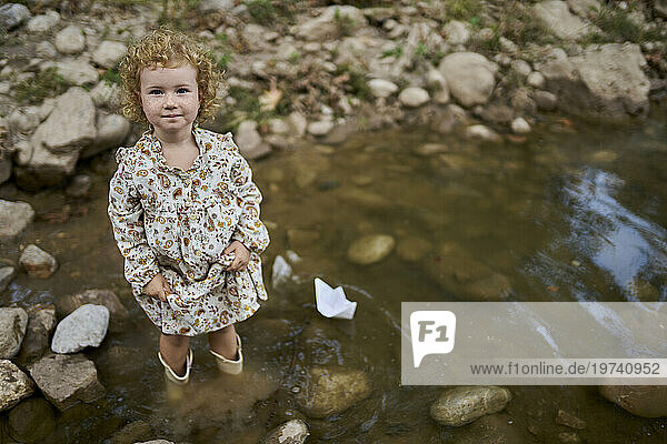 Smiling cute girl standing near paper boat and rocks in water puddle
