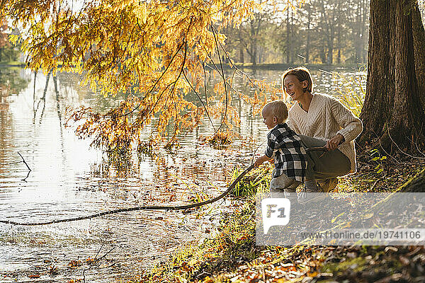 Happy mother with son holding large stick near lake in forest