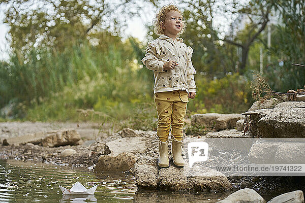 Smiling girl standing on rock with paper boat in water puddle