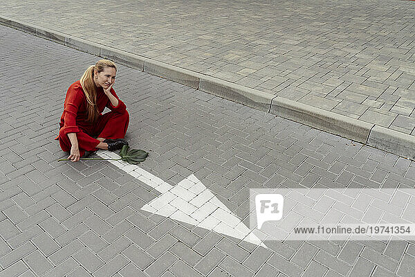 Contemplative woman with monstera leaf sitting near arrow sign on road