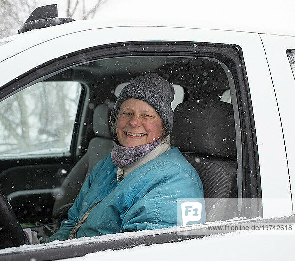 Close-up of a woman farmer sitting in the driver's seat of a car wearing winter clothes and smiling at the camera while snowflakes are falling in winter; Ottawa Valley  Ontario  Canada