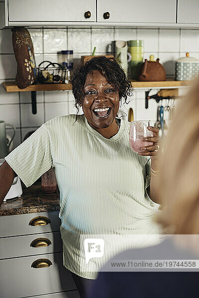 Happy mature woman with smoothie glass talking to female friend at home