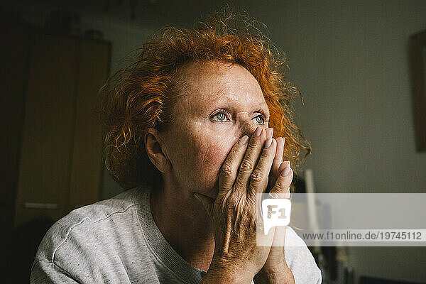 Thoughtful sad senior woman with hands covering mouth sitting at home
