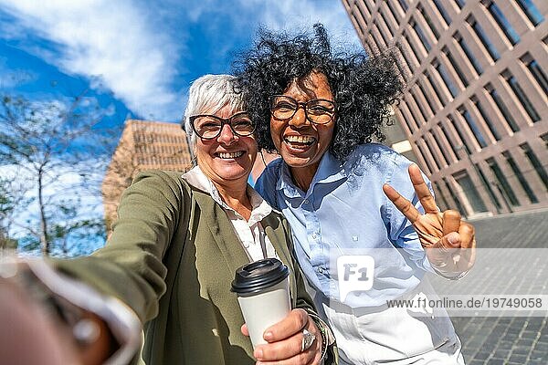 Frontalfoto von zwei multiethnischen lächelnden Geschäftsfrauen  die ein Selfie machen und mit den Fingern im Freien den Sieg gestikulieren