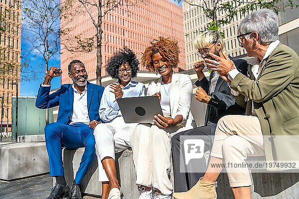 Geschäftsleute  die Nachrichten lächelnd und mit erhobenen Fäusten feiern und dabei einen Laptop im Freien benutzen
