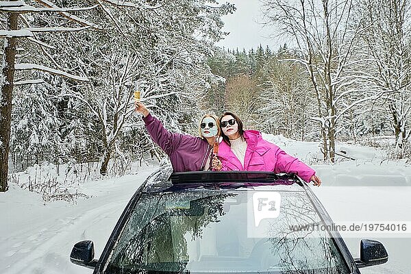 Zwei Frauen mit Gesichtsmasken  die ein Ereignis feiern und sich mit einem Glas Champagner aus der Autoluke lehnen