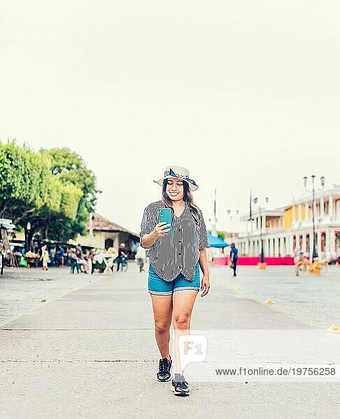 Lächelnder junger Tourist mit Hut und Handy auf der Straße. Weiblicher Tourist texting Telefon auf dem Platz von Granada
