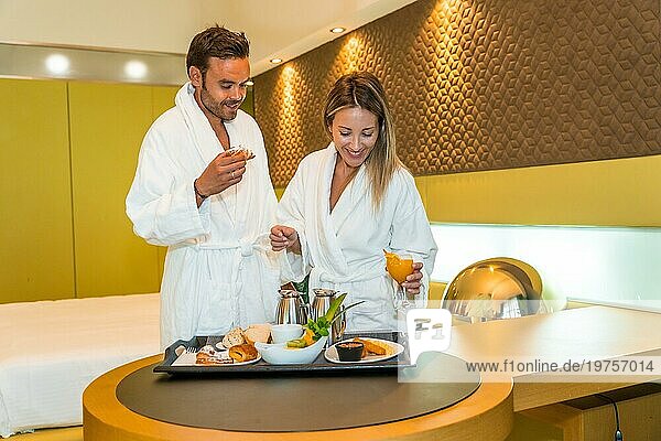 Glückliches frisch verheiratetes Paar im Bademantel beim Frühstück in einem Luxushotelzimmer