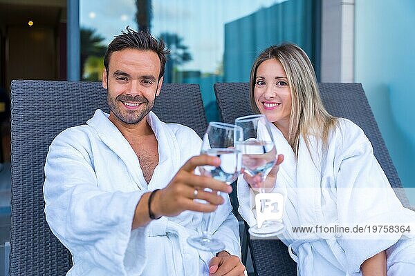 Ein Paar  das seine perfekten Flitterwochen in einem hotelspa feiert  stößt mit Weißwein an  während es in die Kamera schaut und einen weißen Bademantel anzieht  der auf einem Sofa liegt