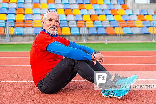 Porträt eines älteren Mannes in Sportkleidung  der auf einer Leichtathletikbahn sitzt und in die Kamera lächelt und müde aussieht