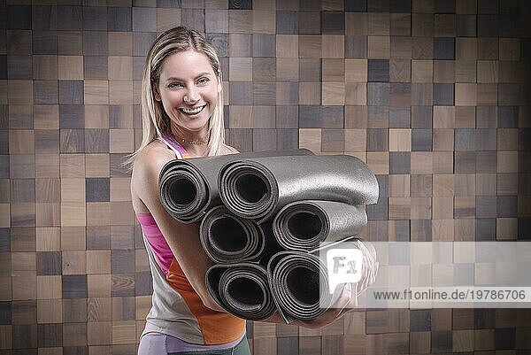 Bild einer fröhlichen erwachsenen Frau  die einen Stapel gerollter Übungsmatten trägt. Yoga und Pilates Konzept. Training  Tanzen und Posieren. Gemischte Medien
