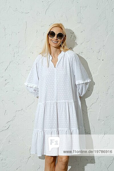 Zufriedene blonde Frau mit Sonnenbrille und weißem Sommerkleid an die Wand gedrückt