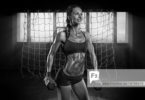 Starke muskulöse Frau posiert in der Turnhalle mit Hanteln in ihren Händen. Das Konzept der Formung  Bodybuilding  Aerobic  Stretching. Gemischte Medien