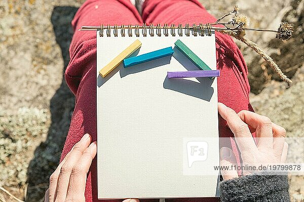 Blick aus der ersten Person auf den weiblichen Schoß ist ein Notizbuch mit Kreiden für die Zeichnung Pastelle. Weibliche Hände halten ein Notizbuch in der Natur