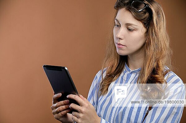 Studioporträt einer attraktiven kaukasischen jungen Studentin in gestreiftem Hemd und Rock mit Hosenträgern. Er hält ein elektronisches Tablet in den Händen und schaut mit einem Lächeln und Freude überrascht auf das Tablet