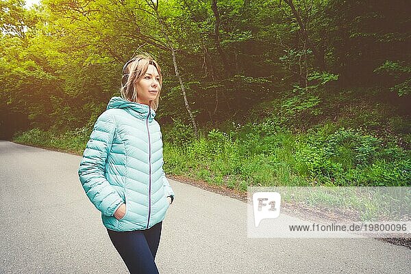 Porträt einer attraktiven  sportlichen Blondine in einer leichten Daunenjacke  die Bluetooth Kopfhörer mit Musik oder Naturgeräuschen anzieht  während sie auf einem Waldweg unterwegs ist