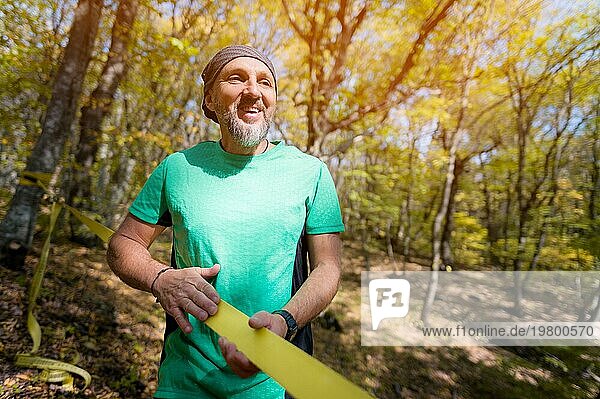 Porträt eines reifen Sportlers mit Bart neben einer gespannten Slackline im herbstlichen Wald am Nachmittag. Das Konzept des Sports nach 40 Jahren