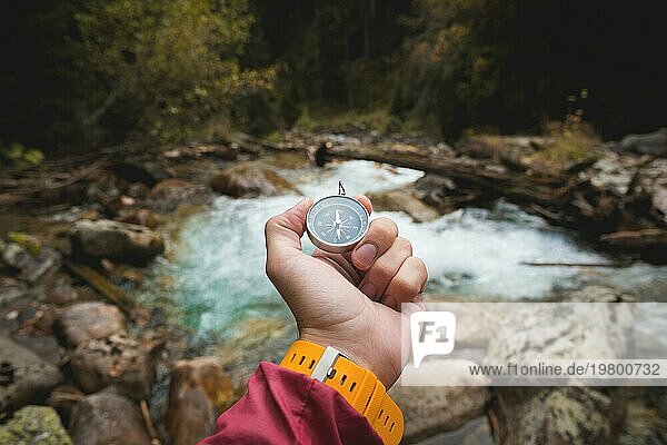 Eine schöne männliche Hand mit einem gelben Uhrenarmband hält einen Magnetkompass in einem herbstlichen Nadelwald vor einem Gebirgsfluss mit felsigen Steinen. Das Konzept  selbst den Weg und die Wahrheit zu finden