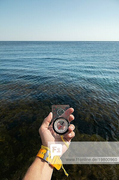 Die Hand eines Mannes mit Armbanduhr hält einen Magnetkompass vor dem Hintergrund des Meeres