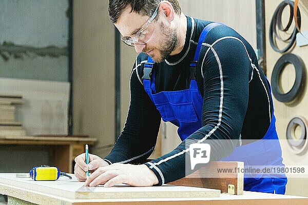 Ein bärtiger  müder  gutaussehender Schreiner sitzt an einem Tisch und zeichnet mit einem Bleistift ein Schild auf die Tafel. Am Arbeitsplatz steht ein gewöhnlicher Meister in blauem Cambenizon mit einem grausamen Haarschnitt und einer gesparten Brille. Das Konzept der Werkstattmöbelproduktion