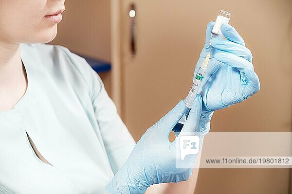 Eine junge kaukasische Ärztin in medizinischer Kleidung und Gummihandschuhen ohne Schutzmaske zieht einen Impfstoff oder ein Medikament aus einer Ampulle in eine Spritze. Nichteinhaltung der medizinischen Sicherheit