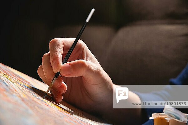Nahaufnahme der Hand einer jungen Künstlerin mit einem dünnen Pinsel  die in einem dunklen Raum ein Bild auf Leinwand malt. Geringe Tiefenschärfe. Hoher Kontrast