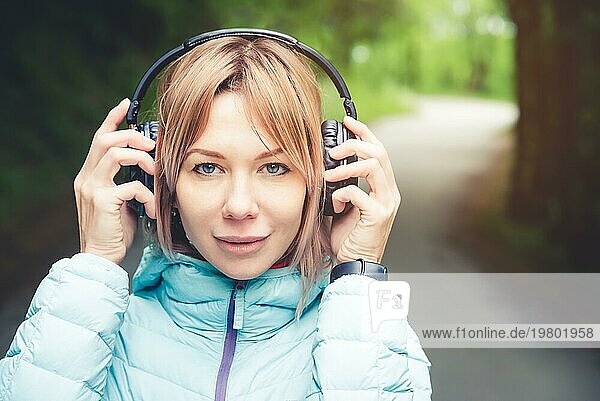Porträt einer attraktiven  sportlichen Blondine in einer leichten Daunenjacke  die Bluetooth Kopfhörer mit Musik oder Naturgeräuschen anzieht  während sie auf einem Waldweg unterwegs ist