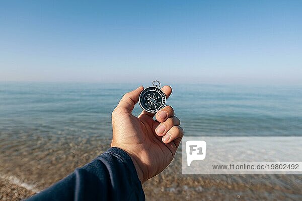 Der Reisende sucht mit einem Kompass an der Küste des morgendlichen Meeres nach dem Weg. Die Hand eines Mannes hält einen magnetischen Kompass