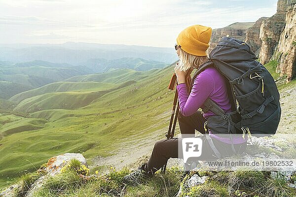 Lächelnde Reisende mit gelbem Hut und Sonnenbrille sitzt am Fuße eines epischen Felsens mit einem Rucksack daneben und schaut weg