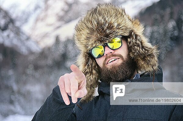 Ein attraktiver bärtiger Mann mit Pelzmütze und Sonnenbrille zeigt mit dem Finger auf die winterlichen Berge im Wald und lächelt