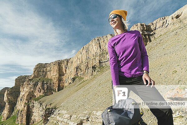 Lächelnde Reisende mit einem gelben Hut und einer Sonnenbrille steht am Fuße eines epischen Felsens mit einem Rucksack daneben und schaut weg