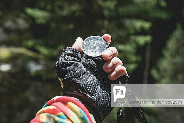 Eine behandschuhte Hand hält einen Magnetkompass vor dem Hintergrund eines Nadelwaldes. Das Konzept der Outdoor Navigation