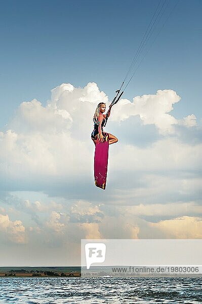 Attraktive schlanke kaukasische junge Frau macht Kiteboarding Stunt gegen Sonnenuntergang Wolken und schönen Himmel. Athlet macht einen Kitesurfing Sprung  während er in die Kamera schaut