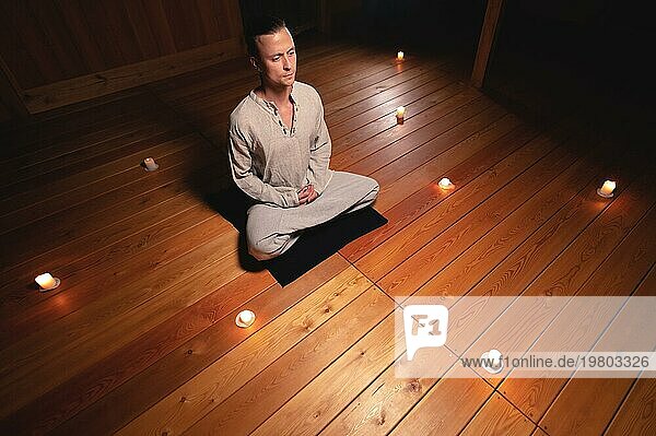 Ein Mann in Praxis und Meditationskleidung sitzt im Lotussitz und hält einen roten Rosenkranz  um seine Aufmerksamkeit in einem hölzernen Raum mit gedämpftem Licht vor dem Hintergrund brennender Kerzen zu konzentrieren. Das Konzept der religiösen Accessoires für Praxis und Aufmerksamkeitsspanne
