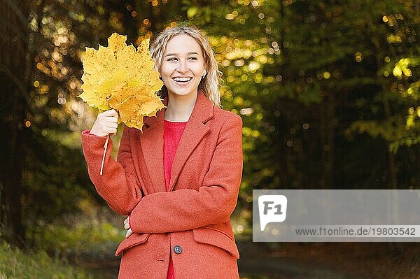 Porträt einer attraktiven jungen Blondine in einem roten Mantel mit einem Strauß gelber Blätter in der Hand. Das Konzept  von Waldspaziergängen während einer Viruspandemie zu träumen. Herbstmode im Wald