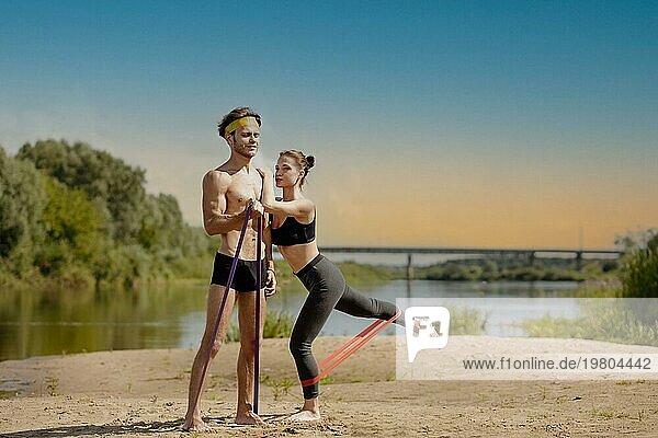 Junges sportliches Paar  das an einem sonnigen Tag am Ufer eines Sees Übungen mit einem Widerstandsband macht