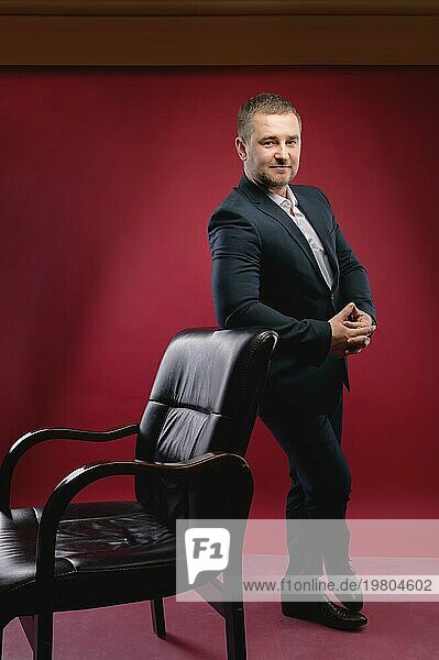 Ein Geschäftsmann mit Bart in einem dunklen Anzug steht neben einem Lederstuhl vor einem burgunderroten Hintergrund  Studioaufnahme