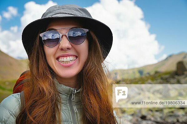 Lächelndes junges Mädchen vor einem blaün  bewölkten Himmel und Bergen. Porträt einer glücklichen Frau Tourist in Sonnenbrille und panama