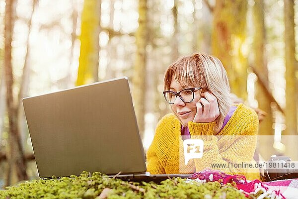 Toned Bild eines Freiberufler Mädchen Porträt in einem gelben Pullover und Brille Blick nachdenklich auf den Laptopbildschirm in der Natur in einem Nadelwald. Das Konzept der Freiberuflichkeit Freiheit für Reisen und das Fehlen der Bindung an das Büro. Ein Laptop liegt auf dem
